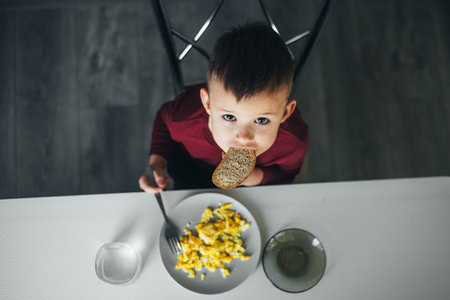 A beautiful little boy in the kitchen is eating a piece of bread, and an omeletの写真素材