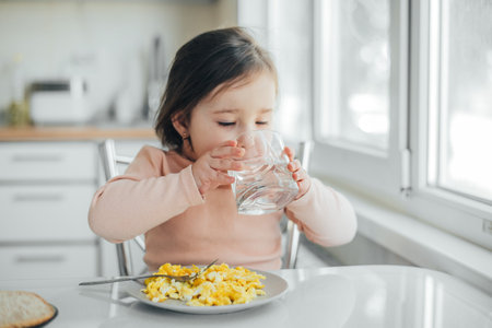 a girl child is in the kitchen drinking water there is a plate with an omelet nextの写真素材