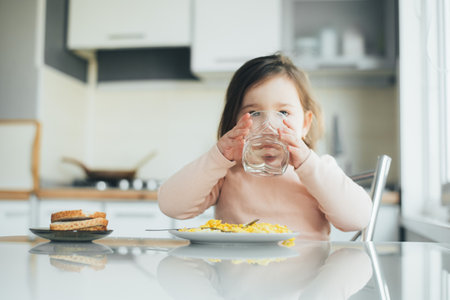 a girl child is in the kitchen drinking water there is a plate with an omelet nextの写真素材