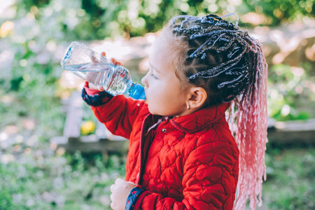 A little beautiful girl with braided pigtails drinks water outdoors in the gardenの写真素材