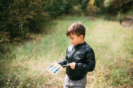 A little boy of five years old is standing in the forest with paints in his hands, trying to paint the leaves of trees with paintsの写真素材