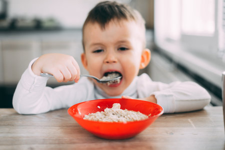 A child in the kitchen eating their own oatmeal with a red plateの写真素材