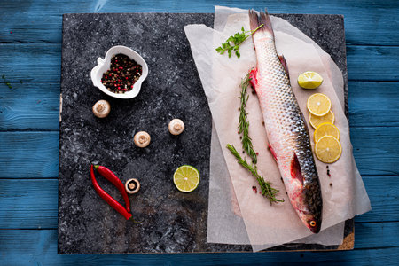 Fresh fish pelengas top view on paper and black granite background , mushrooms , herbs and lime...