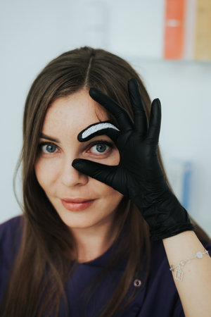 A woman with long brown hair is wearing black gloves and is holding her hand up to her face. She is smiling and she is getting her eyebrows waxedの写真素材