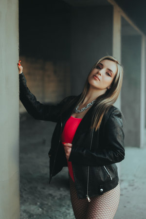 A woman in a red shirt stands in a dark room with a black jacket. She is wearing a silver necklace and has her hands on her hipsの写真素材