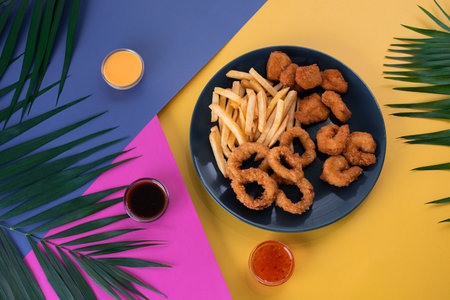 Crispy calamari rings, chicken nuggets, and French fries served on a turquoise plate. Tropical background with palm leaves and colorful saucesの写真素材