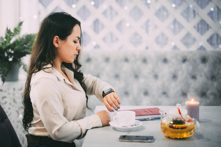 Woman checking her watch while taking a break, seated at a cozy caf table with tea.の写真素材