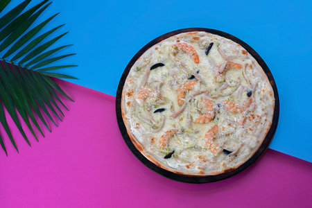 A seafood pizza loaded with shrimp, squid, mussels, and melted cheese, presented on a vibrant blue and pink tropical background with palm leaves.の写真素材