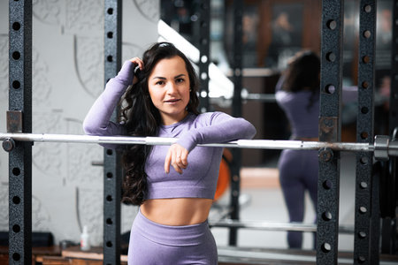 Woman in a purple gym outfit smiling and leaning on a barbell in a gym setting.