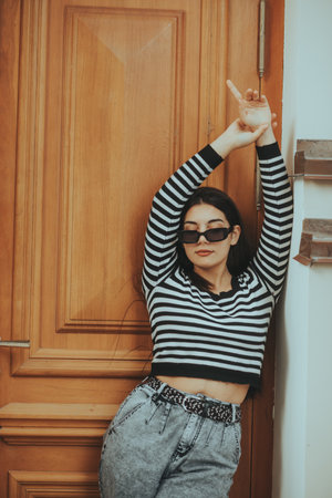 Confident woman in sunglasses poses by a wooden door, embracing modern street style with casual elegance.の写真素材