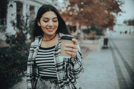 A joyful brunette walks down a city street with a cup of coffee in hand, embracing the autumn air and her stylish street fashion.の写真素材