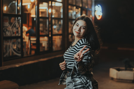 A cheerful brunette strikes a playful pose near a cozy cafe in the autumn night city, surrounded by glowing lights and an urban atmosphere.の写真素材