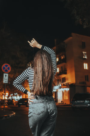 A fashionable brunette with long hair stands with her back to the camera in the autumn night city, arms raised confidently, exuding urban style.の写真素材