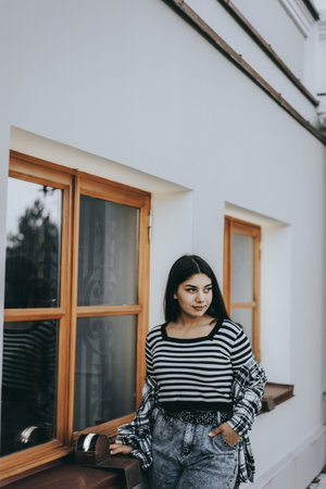 The young woman stands in front of a window, hands on hips, sporting a layered fall outfit.の写真素材