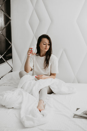 Woman sitting on bed with a drink, relaxed and content, capturing a peaceful morning atmosphere in a cozy bedroom.の写真素材