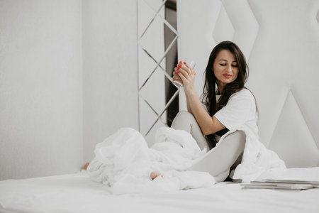 Woman sitting in bed with a drink, smiling and relaxed, creating a positive and cheerful start to her day.の写真素材
