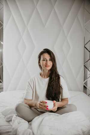 A woman sits on the bed, holding a coffee cup, looking calm and relaxed, setting a peaceful start to her day.の写真素材