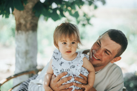 A happy father holds his smiling daughter on his lap, enjoying a warm sunny day in the garden.の写真素材