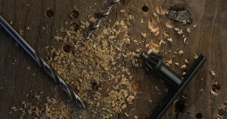 Close-up of drill bits, wood shavings, and a key chuck placed on a weathered wooden table with...