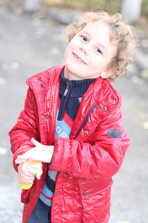 A little curly cheerful boy in a bright red jacket walks on the street in the fall and blowing soap bubblesの写真素材
