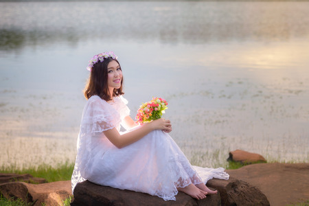 beautiful young woman in white dress with flower on her hair holding flower outdoor shot. Portrait of beautiful blond young woman sitting on rock with water background on sunsetの写真素材