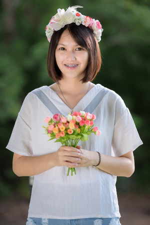 beautiful young woman in white cloths with flower on her hair holding flower outdoor shot. Portrait of beautiful  young woman standing on green tree backgroundの写真素材