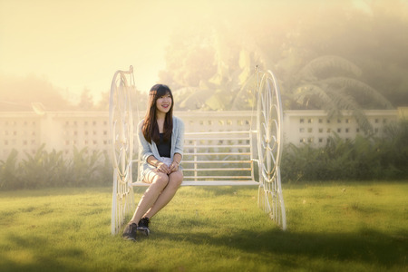 portrait of smiling beautiful Asian woman sitting alone on white bench during sun set with blurry backgroundの写真素材