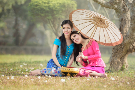 beautiful Asian women (Laos)  in pink and sky blue traditional suit is sitting and smiling happily under Plumeria tree with flower fallen on floorの写真素材