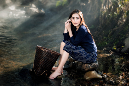 portrait of beautiful Asian woman in traditional costume sitting on stone in creekの写真素材