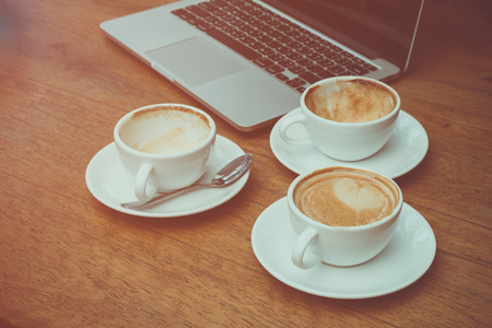 Two empty cups and full coffee cup placing together with laptop on the deskの写真素材