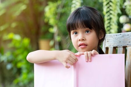 Cute Asian girl looking outside while sitting and reading book in Park