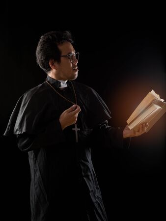 Young Priest with glasses holding old Bible on black background