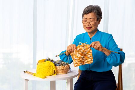 Asian mature senior woman knitting wool clothes and sitting on wooden chair, elderly generation hobby activityの写真素材