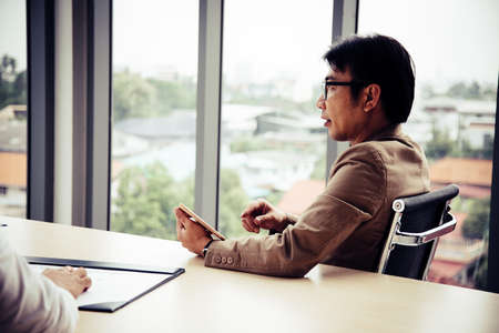Senior manager with glasses holding tablet and looking presentation in meeting roomの写真素材