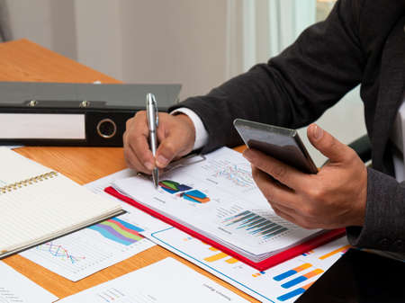 Asian businessman holding check mobile phone with sales data chart sheet