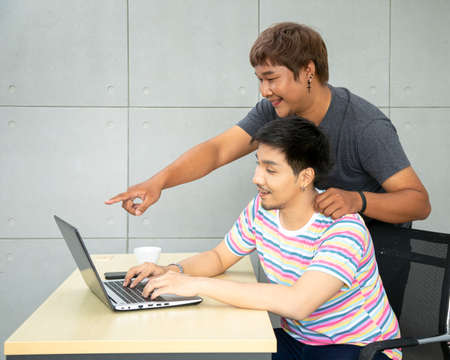 Asian Gay couple at office using internet and laptop shopping online purchase. Fat guy standing pointing displayの写真素材