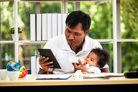 Young father working from home and holding his baby boy feeding milk and digital tablet check information.の写真素材