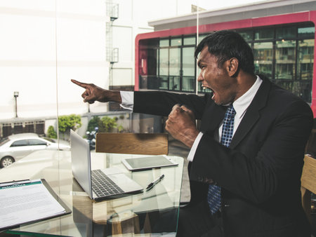 Asian elder businessman in suit and necktie sitting and pointing blame with angry emotion in co-working space cafeの写真素材