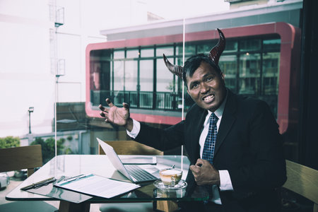 Man in suit with devil horns, expressing aggression in coffee shop with laptop and documents on deskの写真素材