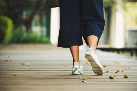 Close-up of person in white sneakers and black pants walking on wooden path in park with fallen...