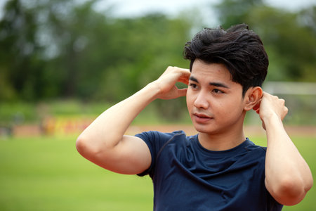 Asian young man in athletic attire relaxes after a workout on a sports field, adjusting his hair and...