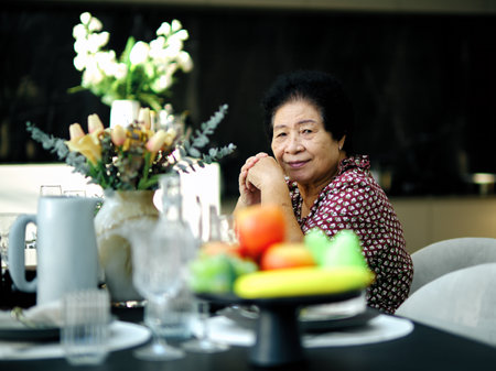 Asian cheerful elderly woman sits at dining table decorated with fruits, flowers, and tablewareの写真素材