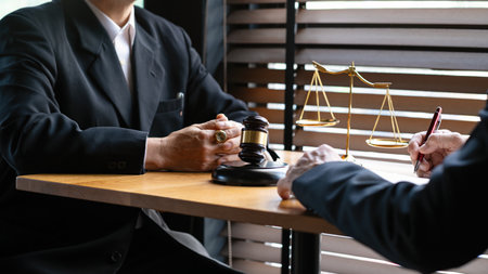 Two lawyers in black suits discussing a legal case at a desk with a gavel, scales of justice, and...