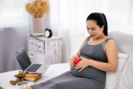 Smiling pregnant woman sits on bed holding a red heart shape on her belly, with laptop, teddy bear,...