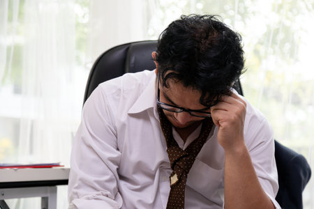 Overwhelmed businessman sitting with head down in office, showing signs of burnout, stress, and workplace fatigueの写真素材