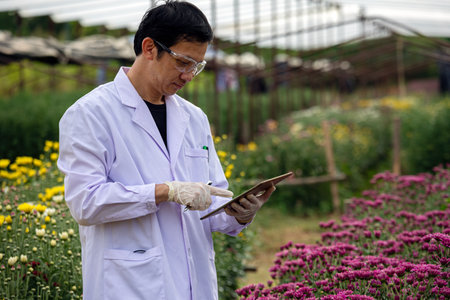 Scientist in white lab coat analyzing flowering plants with a tablet in a greenhouse or field, combining tech and agricultureの写真素材