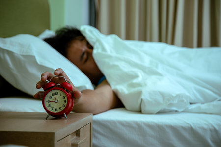 Man lying under white blanket with sleepy face reaching to turn off red alarm clock on wooden nightstandの写真素材
