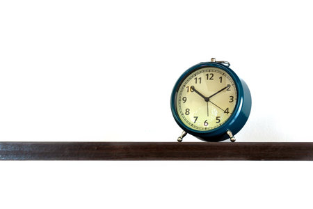 A classic blue alarm clock with metal legs is placed on a dark wooden shelf against a clean white backgroundの写真素材