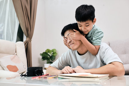 Young child surprises his smiling father by covering his eyes while he works at a table, creating a joyful family momentの写真素材