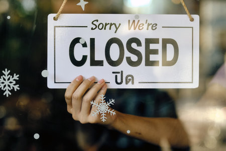 Hand holding a bilingual Sorry Weâ€™re Closed sign on a glass door decorated with winter...